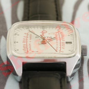 Puede incluir: Un reloj de pulsera plateado vintage con esfera cuadrada y correa de cuero negro. La esfera del reloj es blanca con marcas rojas y la palabra "Raketa". El reloj tiene una corona en el lateral.