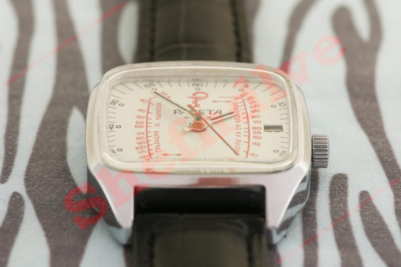 Puede incluir: Un reloj de pulsera plateado vintage con esfera cuadrada y correa de cuero negro. La esfera del reloj es blanca con marcas rojas y la palabra "Raketa". El reloj tiene una corona en el lateral.