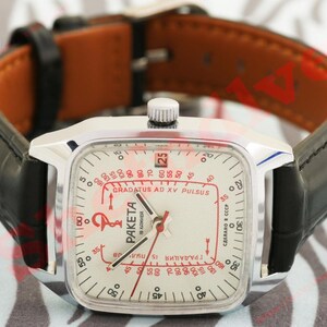 Puede incluir: Un reloj vintage plateado con una esfera cuadrada y una correa de cuero negra. La esfera del reloj es blanca con marcas rojas y negras, incluido el texto "PAKETA" y "GRADATUS AD XV PULSUS". La correa es negra con el interior marr&oacute;n.