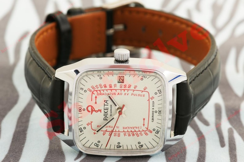 Puede incluir: Un reloj vintage plateado con una esfera cuadrada y una correa de cuero negra. La esfera del reloj es blanca con marcas rojas y negras, incluido el texto "PAKETA" y "GRADATUS AD XV PULSUS". La correa es negra con el interior marr&oacute;n.