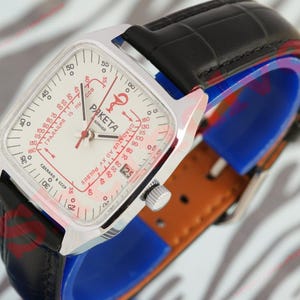 Puede incluir: Un reloj Raketa vintage con una caja cuadrada plateada y una esfera blanca con detalles y texto en rojo. Tiene una correa de cuero negro y un respaldo de cuero marr&oacute;n. La esfera del reloj muestra el nombre de la marca y otras marcas.