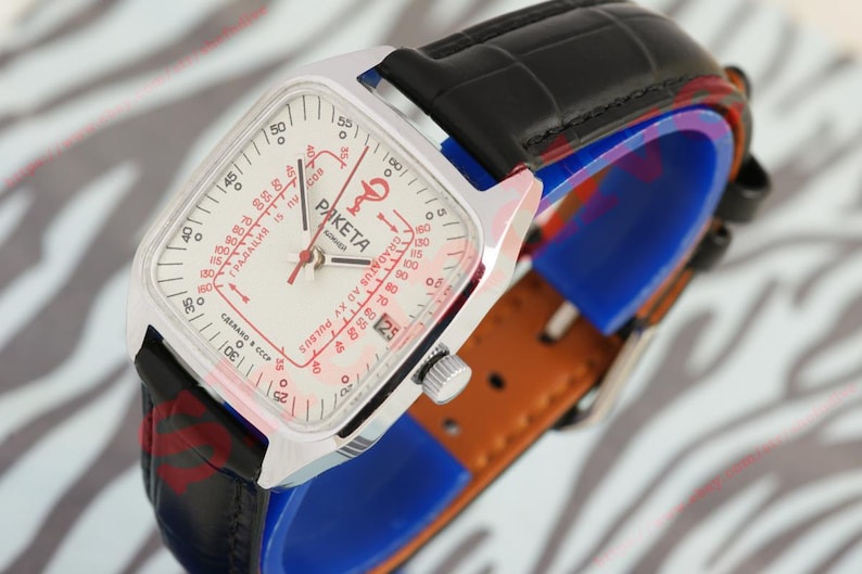 Puede incluir: Un reloj Raketa vintage con una caja cuadrada plateada y una esfera blanca con detalles y texto en rojo. Tiene una correa de cuero negro y un respaldo de cuero marr&oacute;n. La esfera del reloj muestra el nombre de la marca y otras marcas.