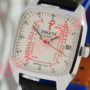 Puede incluir: Un reloj Raketa vintage con caja cuadrada plateada y esfera blanca. La esfera presenta marcas rojas, incluyendo una escala de pulso y la palabra "PAKETA". El reloj tiene una correa de cuero negro y una pantalla de fecha.