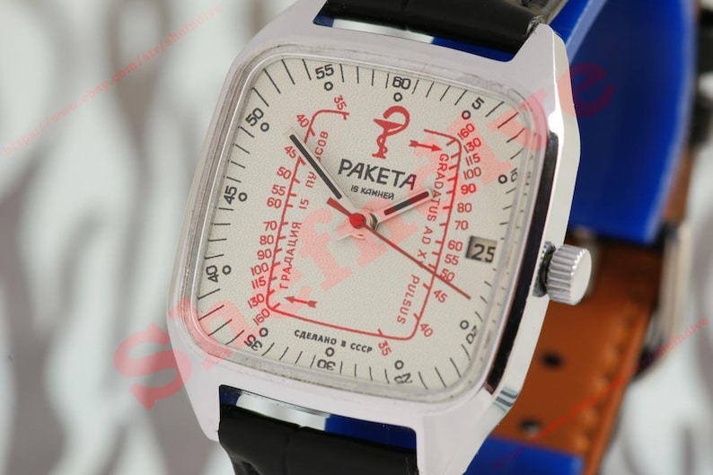 Puede incluir: Un reloj Raketa vintage con caja cuadrada plateada y esfera blanca. La esfera presenta marcas rojas, incluyendo una escala de pulso y la palabra "PAKETA". El reloj tiene una correa de cuero negro y una pantalla de fecha.