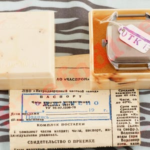 Puede incluir: Reloj de pulsera cuadrado vintage en una caja color crema con documentos. El reloj tiene una caja plateada y una etiqueta que dice "UTK 13". La caja est&aacute; abierta, revelando el reloj y los documentos. El texto est&aacute; en ruso.