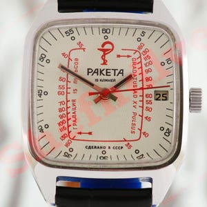 Puede incluir: Un reloj Raketa plateado vintage con esfera cuadrada. La esfera es blanquecina con marcas rojas y negras, incluyendo una escala de pulso y el texto "PAKETA". El reloj tiene una correa negra y una pantalla de fecha.
