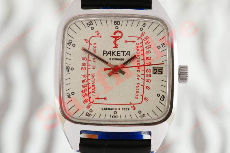 Puede incluir: Un reloj Raketa plateado vintage con esfera cuadrada. La esfera es blanquecina con marcas rojas y negras, incluyendo una escala de pulso y el texto "PAKETA". El reloj tiene una correa negra y una pantalla de fecha.