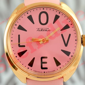 Puede incluir: Un reloj de pulsera rosa con una caja dorada y una correa de cuero rosa. La esfera del reloj tiene marcadores de hora en números romanos negros y una aguja de segundos negra. La esfera del reloj también tiene el texto "Takema" y "Сделано в России" (Hecho en Rusia).