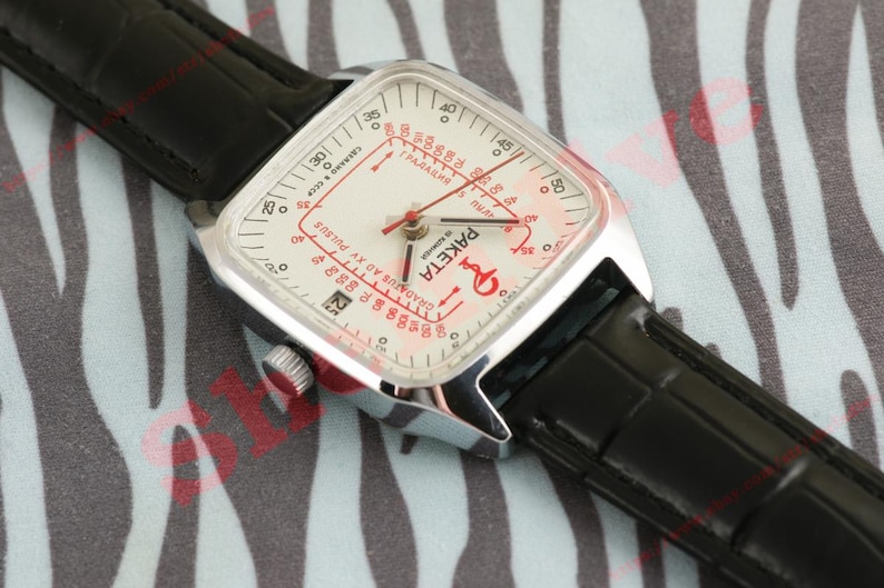 Puede incluir: Un reloj de pulsera plateado vintage con una esfera cuadrada y una correa de cuero negro. La esfera del reloj es blanca con marcas rojas y la palabra "Paketa". El reloj est&aacute; sobre un fondo estampado.