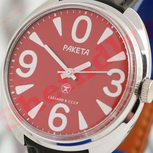 May include: Close-up of a Raketa wristwatch with a red face and white numerals. The watch has a silver-coloured case and a black leather strap. The watch face displays the word "Raketa" and the text "Made in USSR".