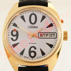 May include: Gold-plated wristwatch with a white face, large black numerals, and black and red triangular markers. The watch has a date display and the word "Слава" is printed on the face. It has a black leather strap.