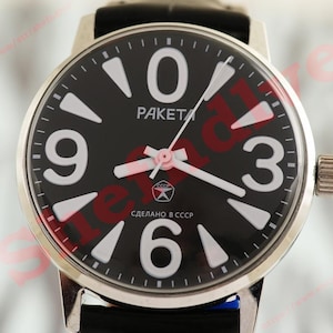 Peut inclure: Une montre analogique noire avec des chiffres et des aiguilles blancs. Le cadran de la montre affiche la marque "Raketa" et du texte en cyrillique. La montre a un boîtier argenté et un bracelet en cuir noir.