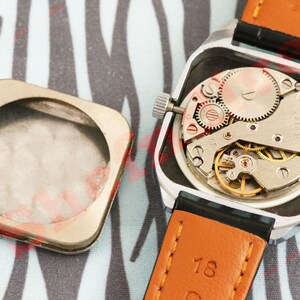 Puede incluir: Un reloj de pulsera vintage con una caja cuadrada plateada, que revela el intrincado movimiento mec&aacute;nico. El reloj tiene una correa de cuero marr&oacute;n con detalles en negro. La parte trasera de la caja del reloj est&aacute; separada.
