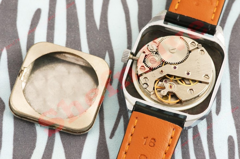 Puede incluir: Un reloj de pulsera vintage con una caja cuadrada plateada, que revela el intrincado movimiento mec&aacute;nico. El reloj tiene una correa de cuero marr&oacute;n con detalles en negro. La parte trasera de la caja del reloj est&aacute; separada.
