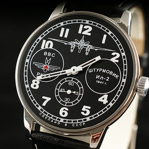 Puede incluir: Un reloj de pulsera negro con caja plateada y correa de cuero negro. La esfera del reloj presenta un gráfico de avión blanco, el texto "ШТУРМОВИК Ил-2 1941 г." y un pequeño subdial con el texto "BBC".