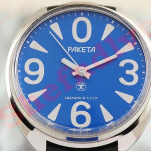 May include: A wristwatch with a blue face, white numerals, and white hands with pink accents. The watch face has the word "PAKETA" and Cyrillic text. The watch has a silver-coloured case and a black strap.
