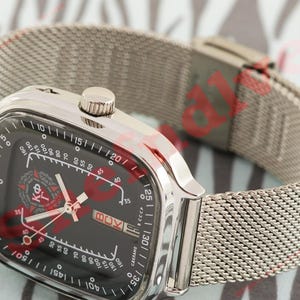 Puede incluir: Un reloj de pulsera plateado con una correa de malla y una esfera negra cuadrada. La esfera del reloj presenta detalles en rojo, marcas blancas y el texto "СДЕЛАНО". El reloj tiene una corona plateada y un cierre.