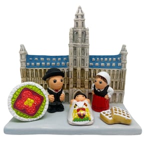 Puede incluir: Un conjunto de figuritas de cerámica que representan a una familia vestida con ropa tradicional belga, de pie frente a una réplica en miniatura de la Grand Place de Bruselas, Bélgica. El conjunto incluye un hombre, una mujer y un niño, junto con una gofre y una flor.