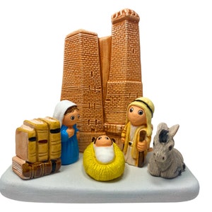 May include: A ceramic nativity scene with a brown building, a baby Jesus in a yellow manger, a donkey, and three figures. The figures are wearing blue, yellow, and white clothing.