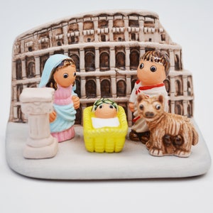 Puede incluir: Un belén de cerámica con un bebé Jesús en un pesebre amarillo, María con un vestido azul y rosa, José con una túnica roja y blanca y un perro marrón. La escena se desarrolla frente a un Coliseo en miniatura.