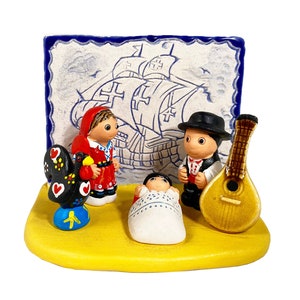 May include: A ceramic nativity scene featuring a baby Jesus, Mary, Joseph, a black rooster, and a man playing a guitar. The scene is set against a blue and white tile depicting a ship.