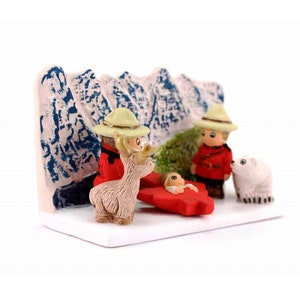 Puede incluir: Una escena de la natividad navide&ntilde;a con una hoja de arce roja, un oso polar blanco, un reno marr&oacute;n y dos figuras vestidas con uniformes de guardabosques rojos y verdes. La escena est&aacute; ambientada en un fondo blanco con una cordillera azul y blanca.