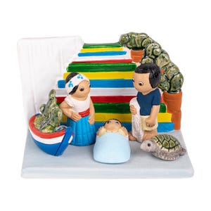 May include: A ceramic nativity scene with a blue and white striped staircase, a green cactus, a blue boat, a green turtle, and three figures: a woman in a blue dress, a man in a blue shirt, and a baby in a blue blanket.