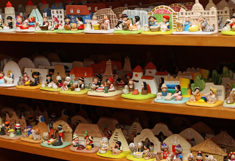 Puede incluir: Una colecci&oacute;n de figuritas en miniatura que representan diversas escenas y personajes. Las figuritas est&aacute;n hechas de cer&aacute;mica pintada y est&aacute;n dispuestas en estantes. Las figuritas incluyen una escena de la natividad, un grupo de ni&ntilde;os jugando y un grupo de personas con ropa tradicional.
