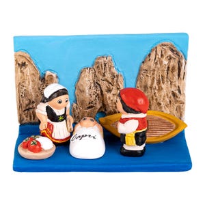 Op de afbeelding: Een keramische figuurtjesset die een kerststal voorstelt met een baby in een witte doek, een vrouw in een rood-witte jurk en een man met een rode hoed en een wit overhemd. De scène speelt zich af tegen een blauwe achtergrond met bruine rotsformaties. De baby is gelabeld met "Capri".