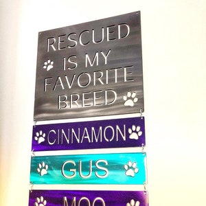 May include: Metal sign with the text "Rescued is my favorite breed" and the names "Cinnamon", "Gus", and "Moo" on separate lines below. Each line has a paw print on either side.