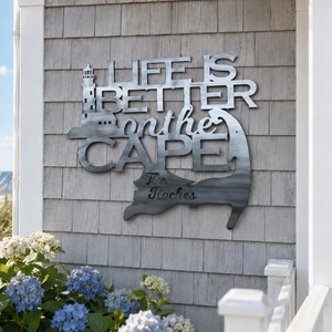Könnte beinhalten: Ein silberfarbenes Metall-Wandschild mit einer Leuchtturm-Silhouette und dem Schriftzug "LIFE IS BETTER on the CAPE" sowie dem eingravierten personalisierten Namen "The Roches", geformt nach dem geografischen Umriss von Cape Cod, Massachusetts.