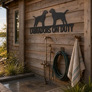 May include: A black metal wall sign featuring silhouettes of two Labrador dogs and the text "LABRADORS ON DUTY" mounted on a wooden exterior wall above an outdoor shower fixture, a coiled green garden hose, and a hanging beige striped towel.