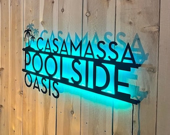 May include: A black metal wall sign featuring the text CASAMASSA POOLSIDE OASIS with a palm tree graphic. The sign is illuminated from behind with a bright turquoise LED light, casting a vibrant glow against the surface behind the metal lettering.