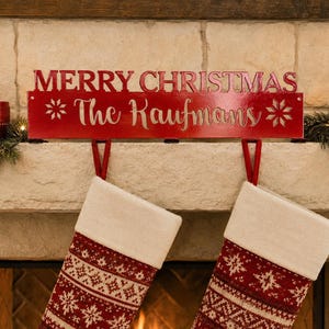 May include: A red metal stocking holder featuring the text MERRY CHRISTMAS and The Kaufmans in white script. The rectangular sign includes two snowflake icons and two red hooks for hanging stockings, designed for placement on a fireplace mantel.