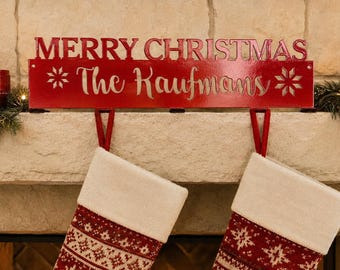 Op de afbeelding: Een rode metalen kerstsokhouder met de tekst MERRY CHRISTMAS en The Kaufmans in witte schrijfletters. Het rechthoekige bord bevat twee sneeuwvlokpictogrammen en twee rode haken voor het ophangen van kerstsokken aan een schoorsteenmantel.
