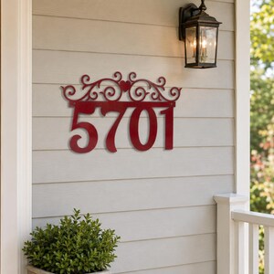 May include: A dark red metal house number sign displaying the digits 5701. The sign features a decorative top border with intricate scrollwork and a small heart shape centered above the numbers, mounted horizontally on a light-colored wall.