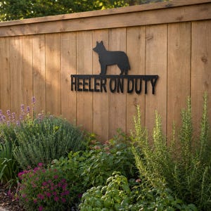 May include: A black metal sign featuring a silhouette of a dog standing above the text HEELER ON DUTY. The sign is mounted on a vertical wooden fence panel, displaying a clean, flat design with sharp edges and a matte finish for outdoor display.