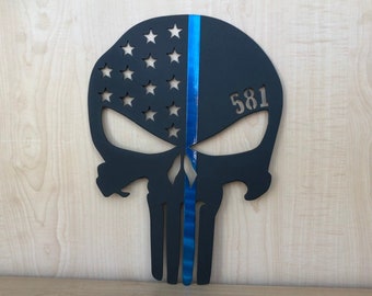May include: A black skull-shaped wall decoration featuring a cutout American flag pattern with stars on the left side and a vertical metallic blue stripe running down the center. The number 581 is cut out on the right side of the skull forehead.