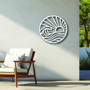 May include: A white, circular wall art piece depicting a sunrise over ocean waves. The artwork is mounted on a white wall next to a wooden chair with a white cushion and pillow. The scene is outdoors with greenery.