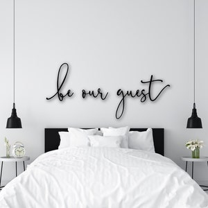 Peut inclure: Chambre moderne avec lit blanc, literie et oreillers. Le mur au-dessus du lit affiche l'inscription "be our guest" en noir. Des suspensions noires sont de chaque côté du lit.