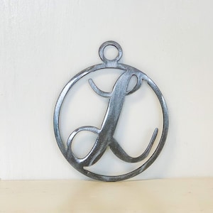 May include: A metal ornament featuring the letter "L" in a cursive script, enclosed within a circular frame. The ornament has a loop at the top for hanging and is made of a silver-colored metal.