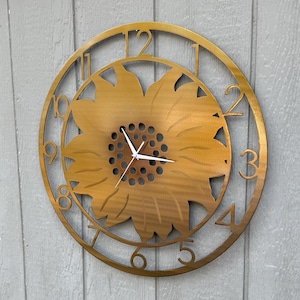 May include: A golden metal wall clock featuring a sunflower design. The clock has a circular frame with numbers and a sunflower in the center. The clock hands are white. The clock is mounted on a light gray wooden wall.