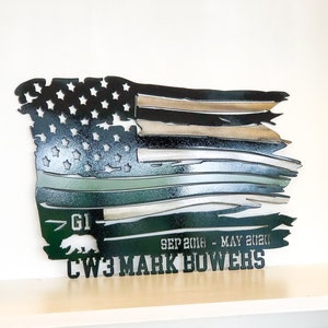 May include: A distressed American flag metal wall art with a green camouflage stripe, a bear silhouette, and the text "GI", "SEP 2016 - MAY 2020", and "CW3 MARK BOWERS".