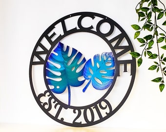 Op de afbeelding: Zwart metalen welkom bord met een cirkelvormig ontwerp en de woorden "Welcome Est. 2019". Twee blauwe en groene tropische bladeren bevinden zich in de cirkel.
