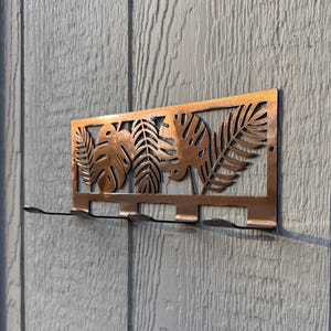 May include: A copper-colored metal wall hook with a tropical leaf design. The rectangular piece features cut-out monstera and fern leaves. Below the leaves are four hooks for hanging items. The hook is mounted on a light gray wooden wall.