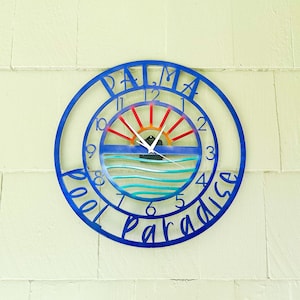 Może przedstawiać: Metalowy zegar w kolorze niebieskim i białym z napisem "Palma Pool Paradise" wokół krawędzi. Tarcza zegara przedstawia kolorowy wschód słońca i fale oceanu.