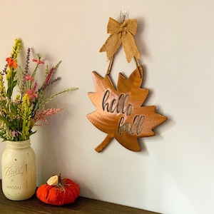 May include: A wooden leaf-shaped sign with the text "hello fall" hanging on a white wall. A bouquet of flowers in a white mason jar and a small orange pumpkin are on a wooden table below the sign.