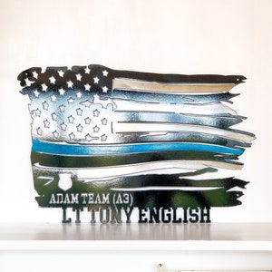 May include: A distressed American flag metal wall art with a thin blue line and the text "Adam Team (A3) LT Tony English".