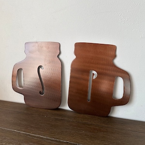 May include: Two metal mason jar silhouettes with the letters "SP" cut out. The jars are a copper color and are mounted on a wooden surface.