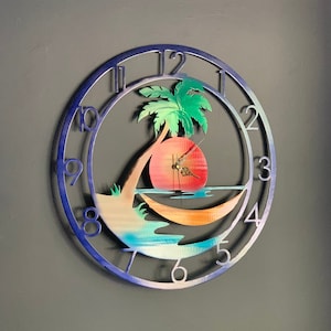 Puede incluir: Un reloj de pared de metal azul con un diseño de palmera y puesta de sol. La esfera del reloj tiene números romanos y un sol rojo con una manecilla de reloj.